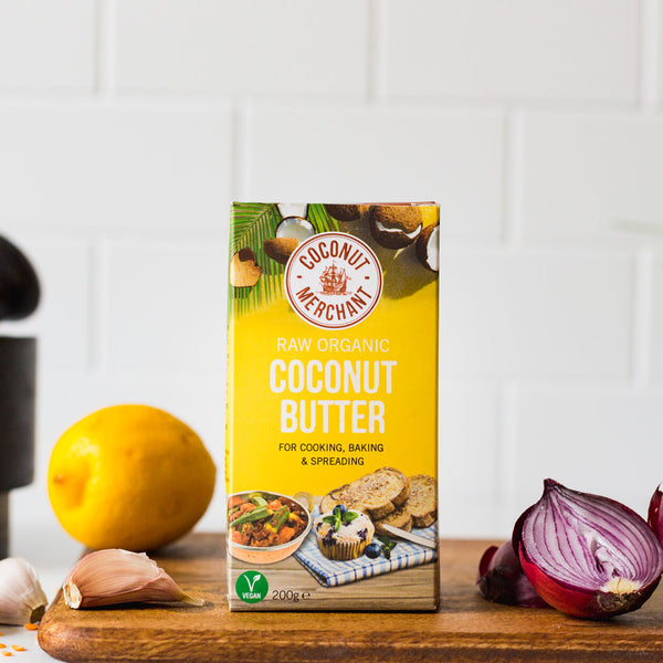 Coconut Merchant Raw Organic Coconut Butter 200g on a wooden board with garlic, onion, and lemon in a kitchen setting.