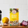Coconut Merchant Raw Organic Coconut Butter 200g on a wooden board with garlic, onion, and lemon in a kitchen setting.