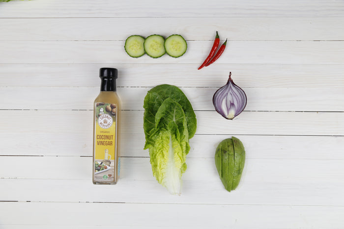 Organic Coconut Vinegar 500ml with fresh salad ingredients including avocado lettuce and cucumber on white wooden background